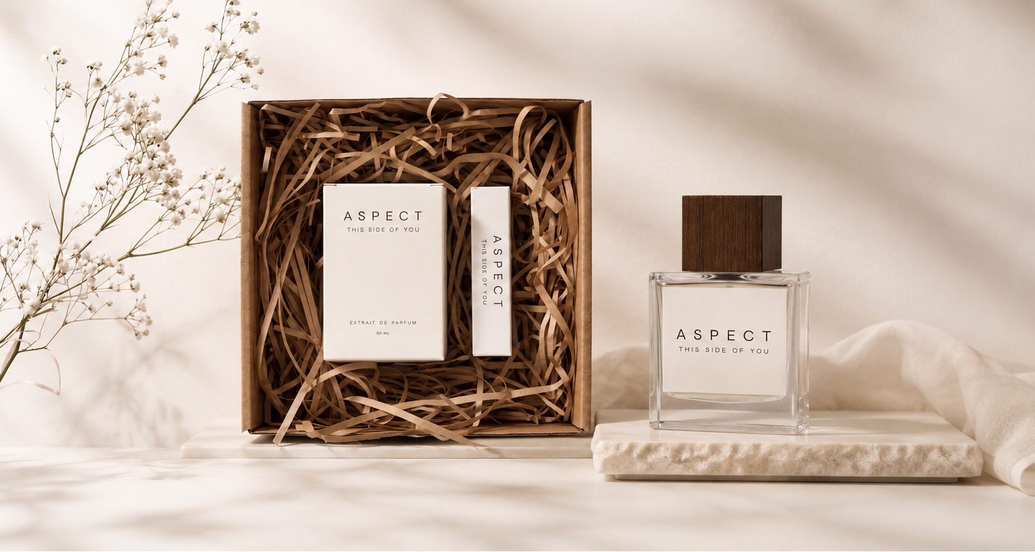A fragrance gift set from Aspect, featuring a clear glass bottle with a dark wood cap and a matching boxed set with a white card and stick, arranged on a cream fabric surface with dried baby's breath flowers and soft shadows.