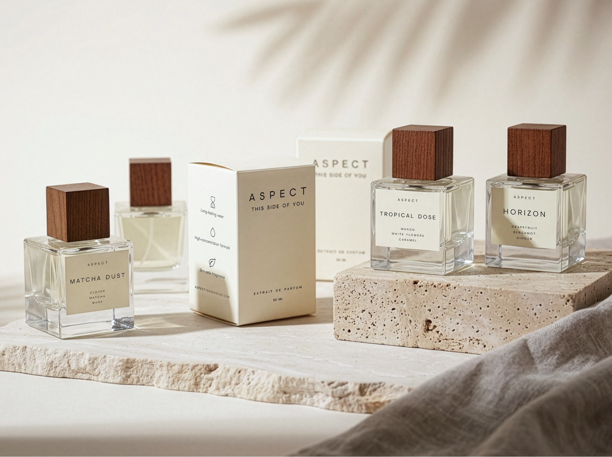 A set of clear glass perfume bottles with dark wood caps and cream-colored boxes, arranged on a stone slab with a draped fabric and soft leaf shadows in the background.