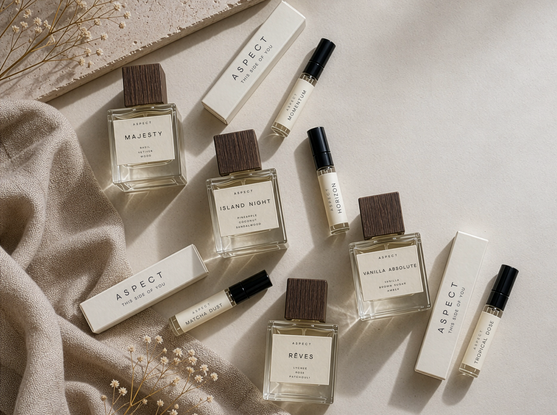 A styled flat-lay of multiple perfume bottles by ASPECT fragrances, including cubic glass bottles with dark wood-grain caps and rollerball bottles, arranged on a light neutral surface with draped beige fabric and dried flowers.